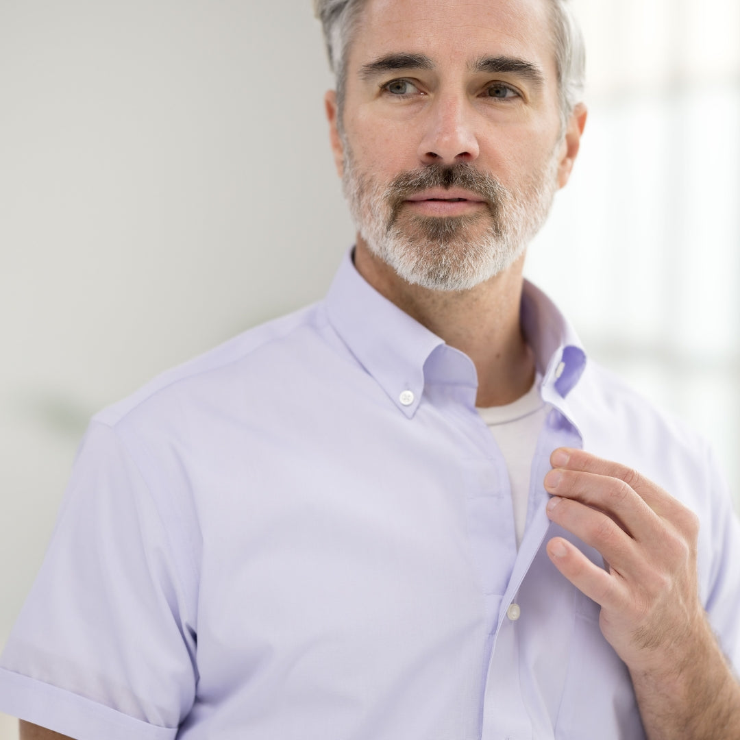 Man in a light purple short-sleeve shirt, adjusting his collar in a bright indoor setting.