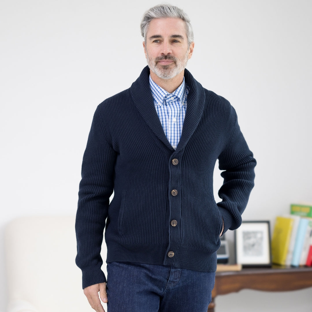Man in navy ribbed cardigan and checkered shirt, posing confidently in a bright, modern living space.