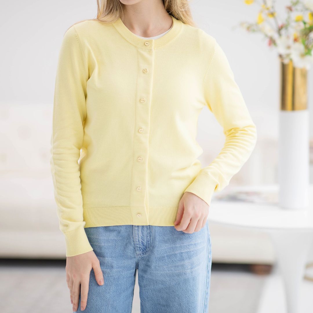 A woman wearing a yellow cardigan with buttons, paired with blue jeans, standing in a bright, airy room.