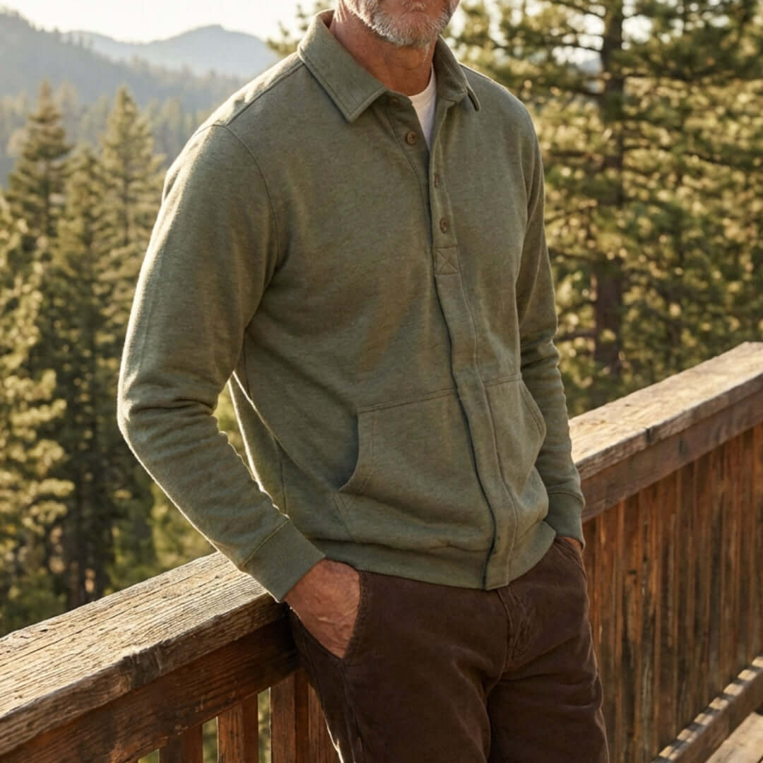 The Adaptive Magnetic Front Sweatshirt in Heathered Olive worn by a man standing on a wooden deck surrounded by trees.