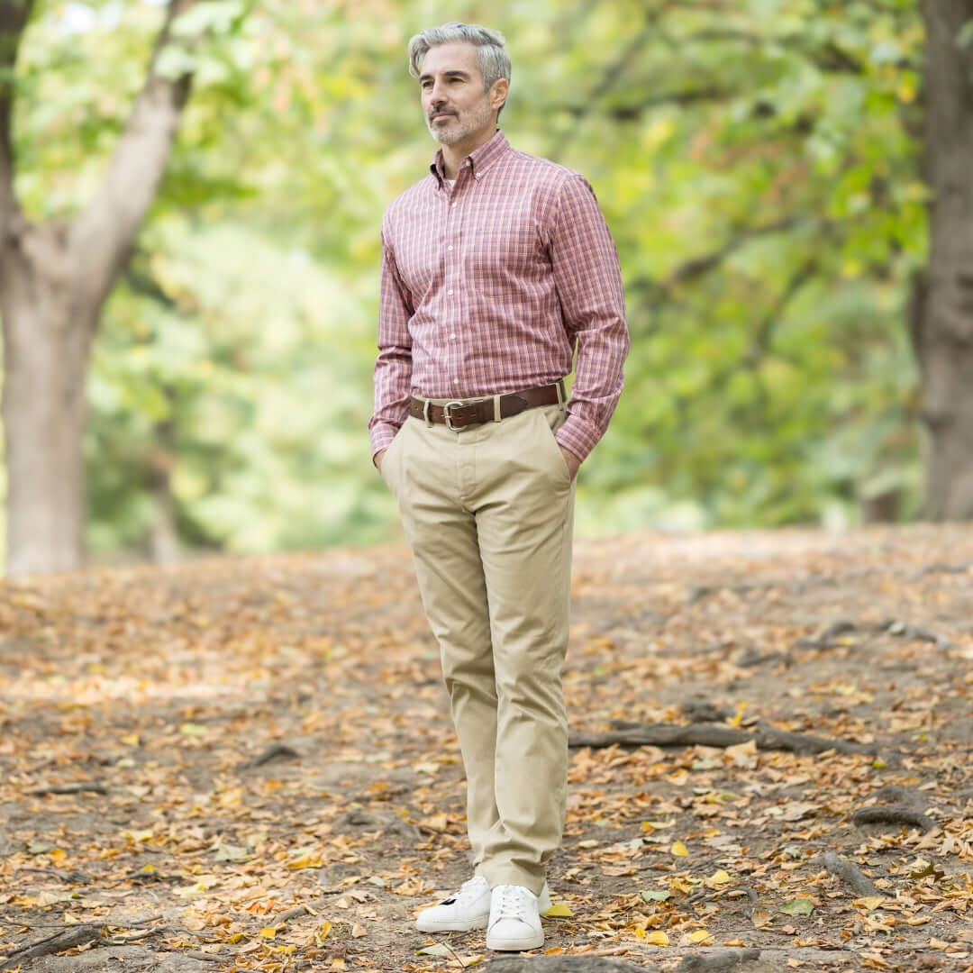 Man in a red checked shirt and beige pants standing outdoors in a park with fallen leaves.