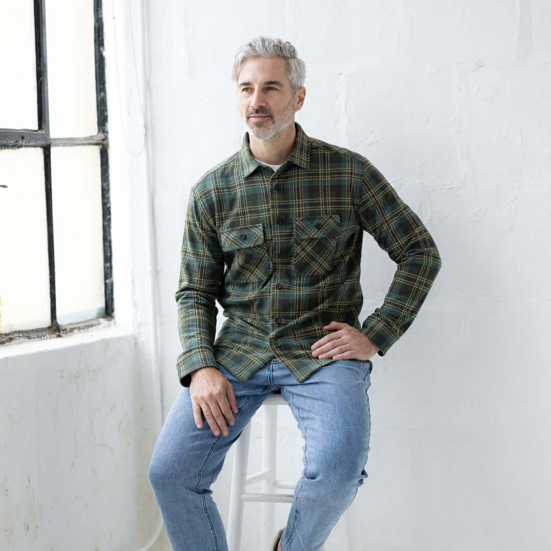 Adaptive Olive Plaid Flannel Shirt with Magnetic Closures worn by a man sitting on a stool in a bright room.