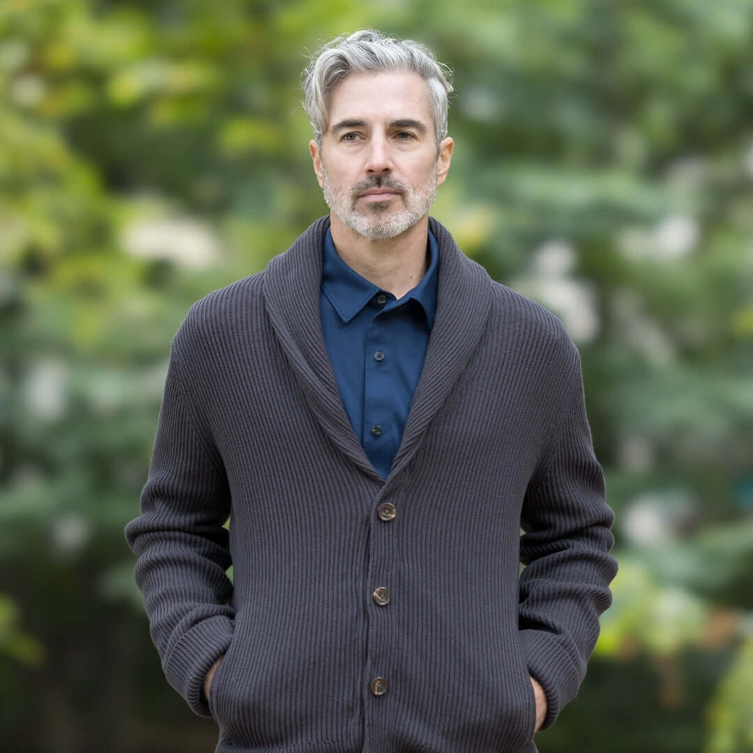 Man wearing a dark cardigan over a navy polo shirt, standing outdoors in a blurred natural setting.