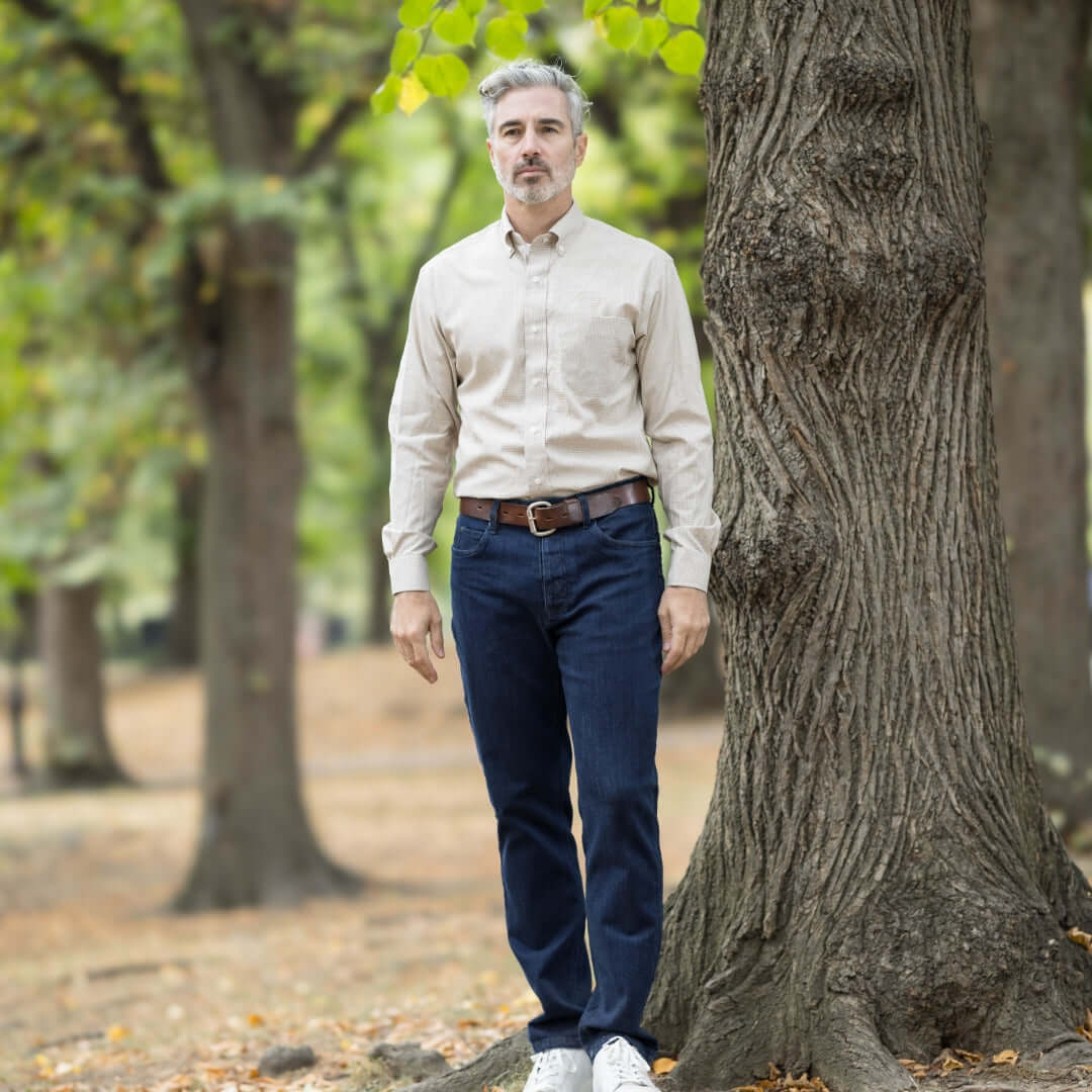 Man standing by a tree wearing light beige shirt and navy blue jeans in a park setting.