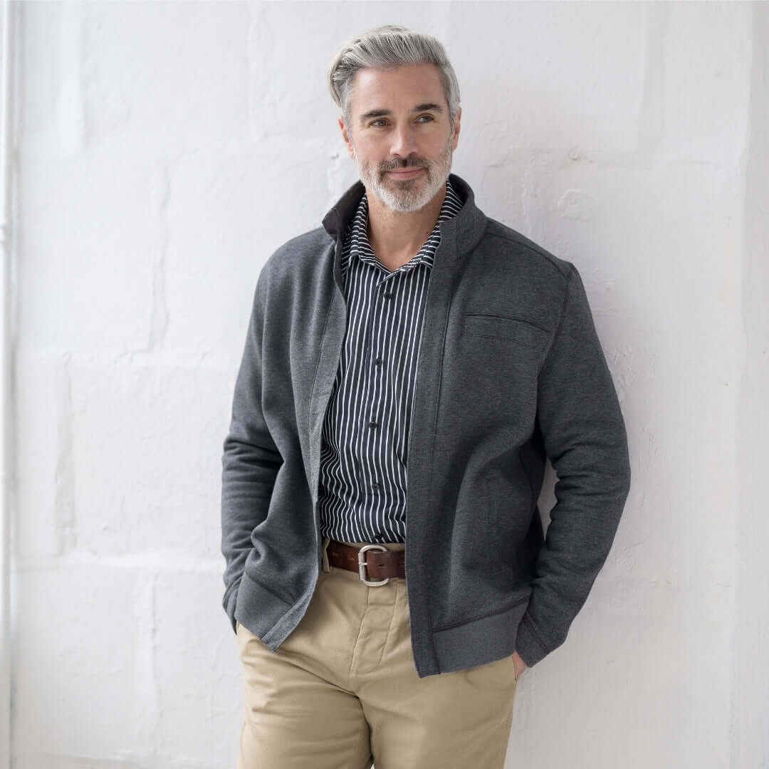 Man wearing a gray zip-up jacket and striped shirt, posing casually against a white wall.