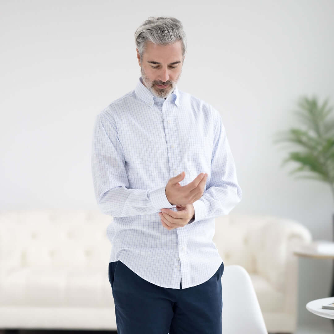 Man adjusting sleeve of navy grid long sleeve button-down shirt in a bright living space.