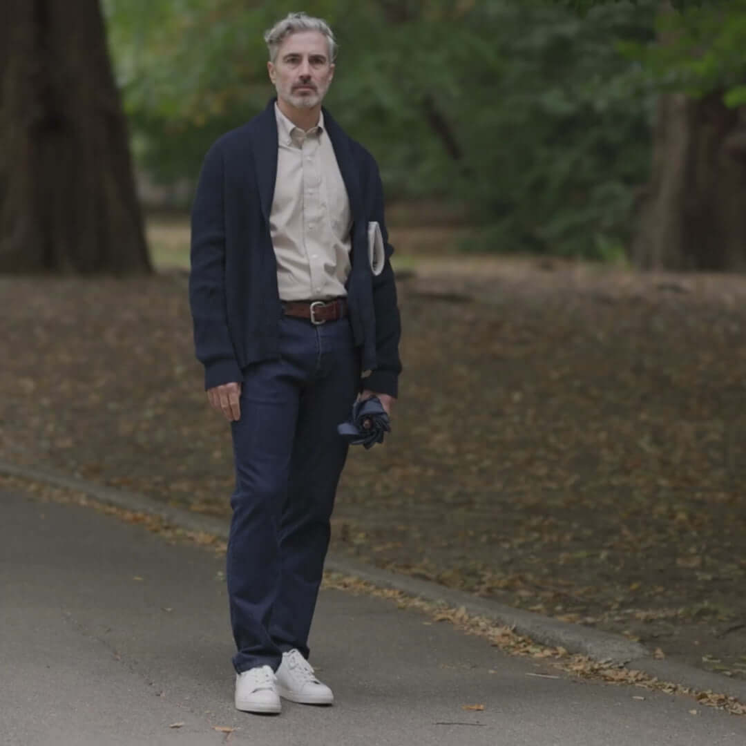 Man wearing a navy ribbed cardigan, beige shirt, and jeans, standing on a park path surrounded by trees.