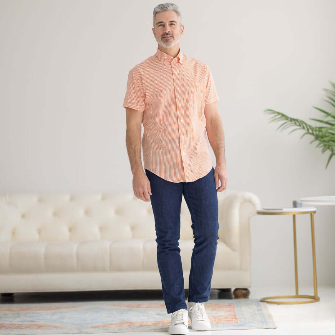 Man wearing a short sleeve orange and white gingham cotton shirt with blue jeans, standing in a bright room.