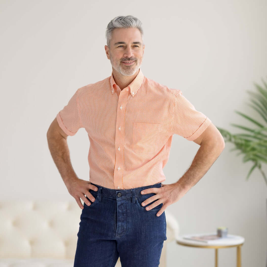 Man in short sleeve orange and white gingham shirt, smiling and standing in a bright room with palm plant.