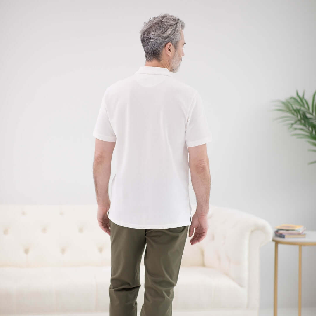 Back view of a man wearing a white pique knit polo shirt with short sleeves and magnetic closures, sitting in a bright room.