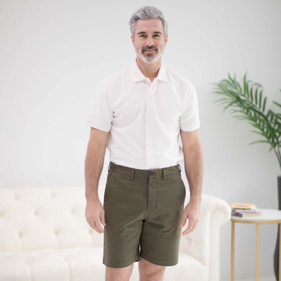 Man wearing a white short sleeve polo and olive shorts, standing in a stylish living room with a sofa and green plant.