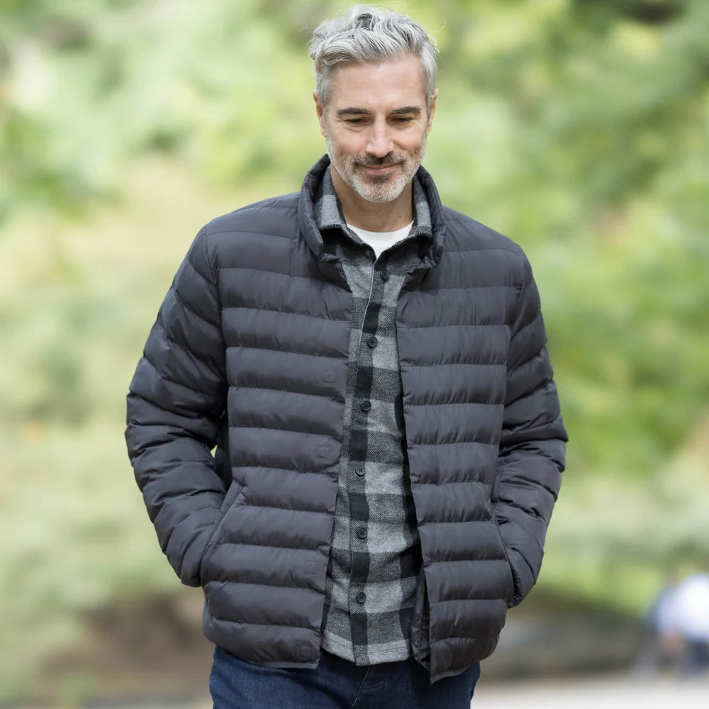 Stylish man wearing a black puffer jacket and plaid shirt, enjoying a walk in a green outdoor setting.