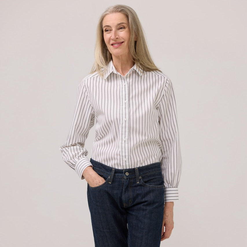 A woman modeling a white long-sleeve button-up shirt with thin vertical black and white stripes, paired with dark blue jeans, against a plain light gray studio background.