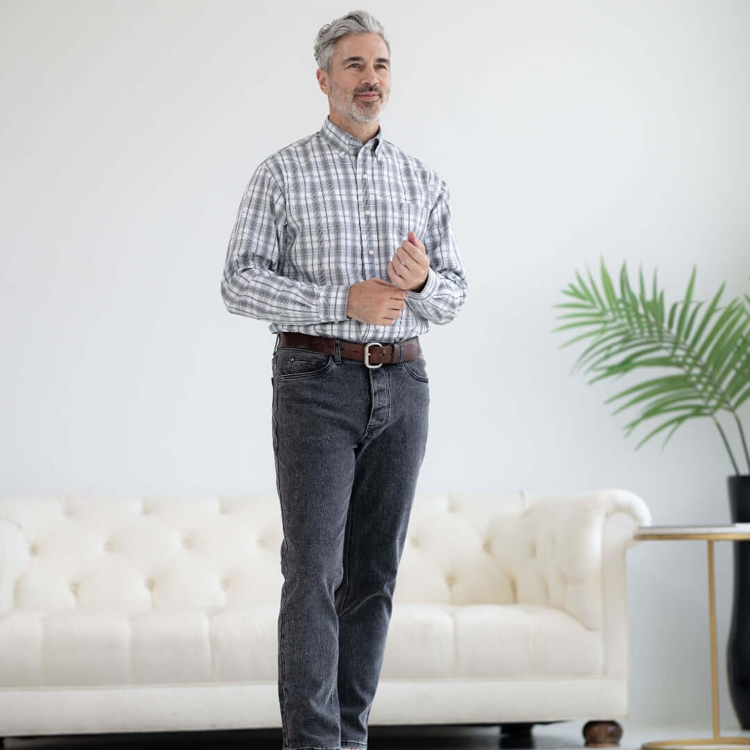 Man in a checkered shirt and black wash jeans standing in a modern living room with a cozy sofa.
