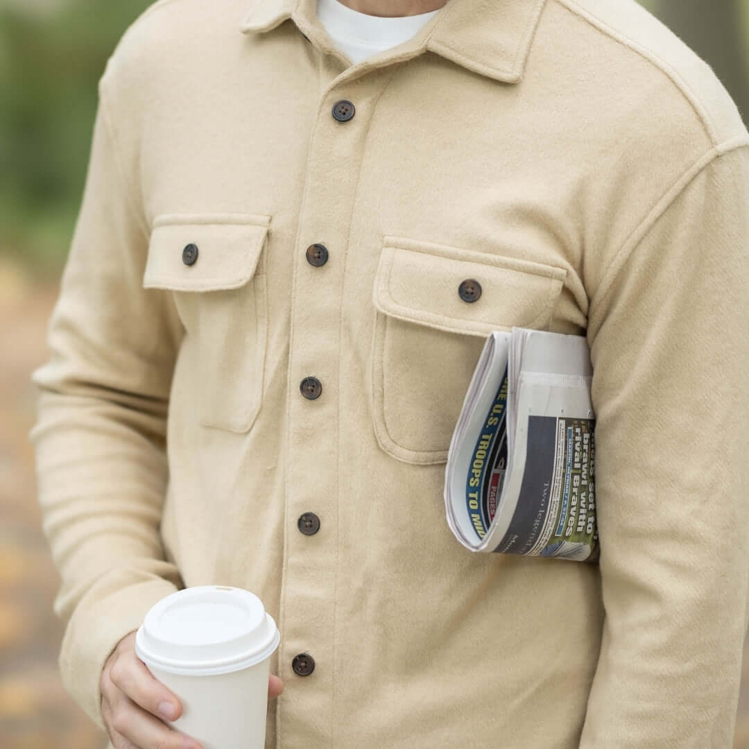 Man in khaki flannel shirt holding a coffee cup and a newspaper, showcasing a stylish layering piece.