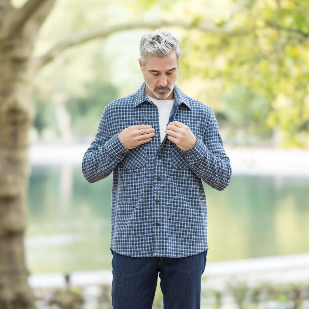 Man adjusting buttons on a stylish white and navy check flannel shirt by a serene lake in autumn.