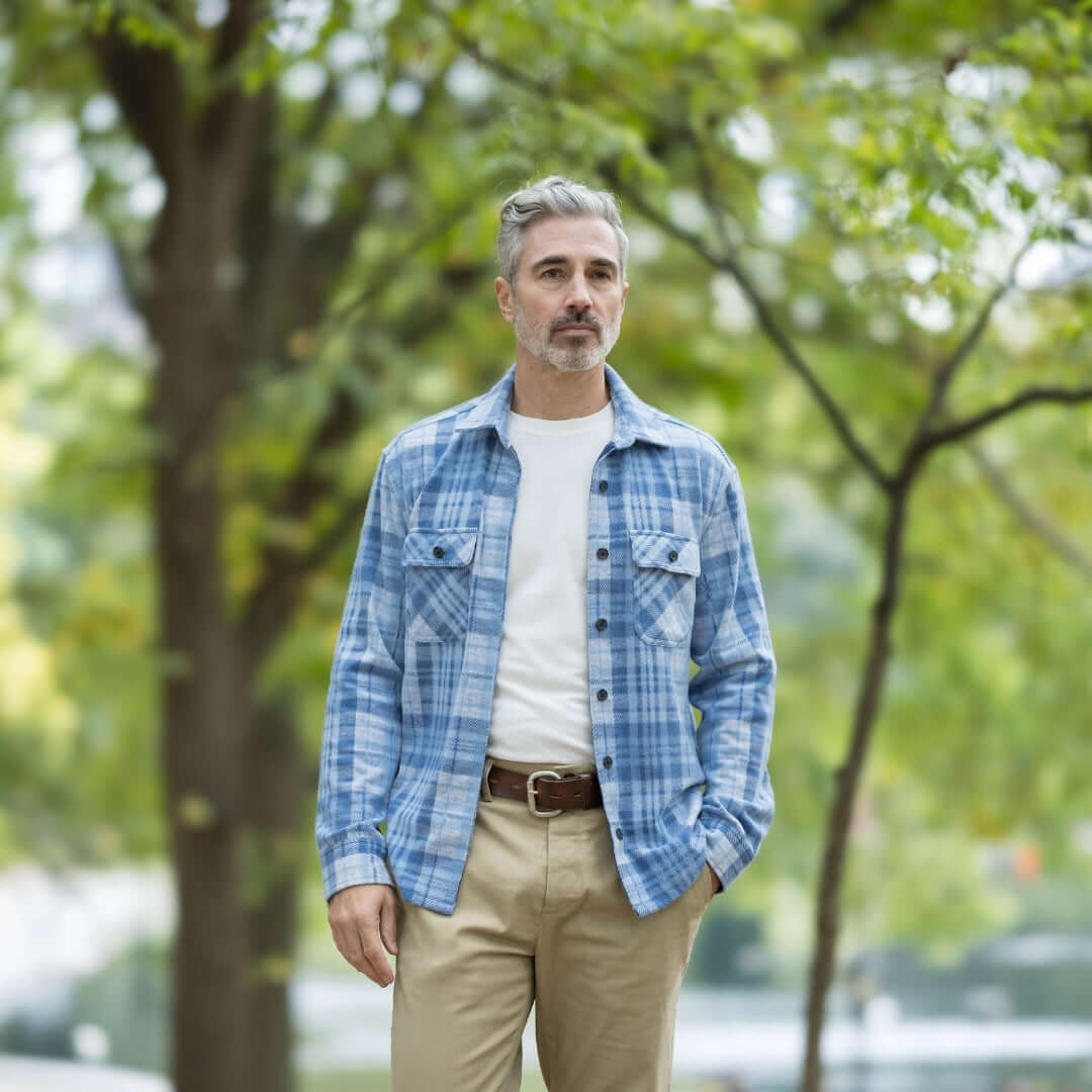 Man wearing pale blue plaid flannel shirt and beige pants, walking outdoors in a park.