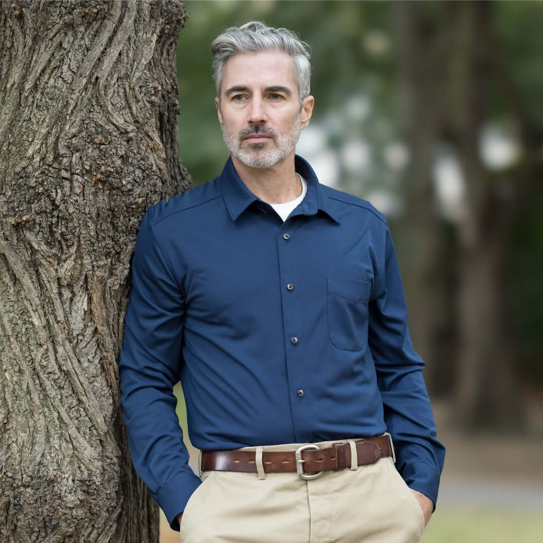 Man wearing adaptive true navy magnetic long sleeve cooling tech polo with pocket, leaning against tree.