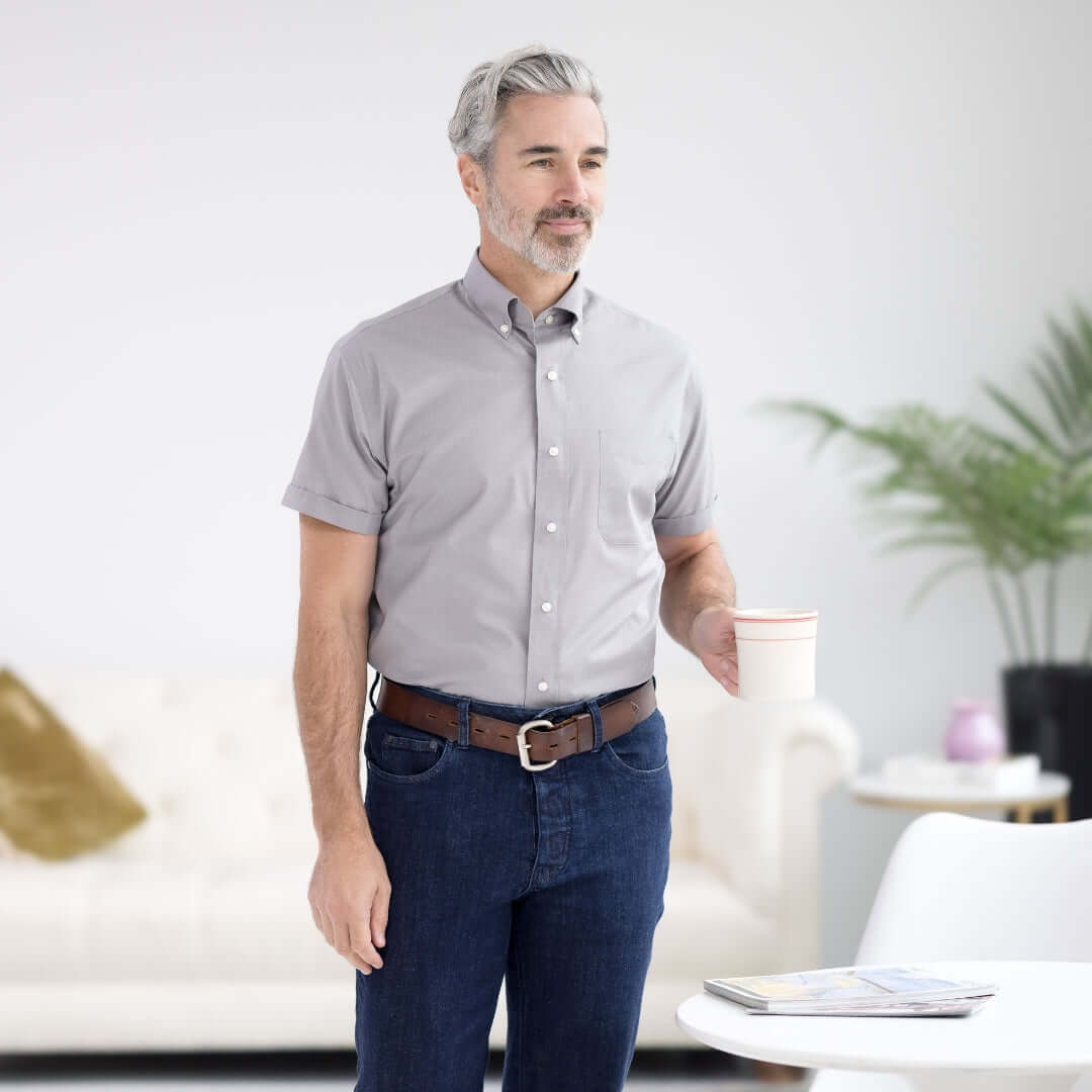 Man wearing a Dove Grey Short Sleeve Button Down Shirt with Magnetic Closures, holding a cup in a stylish living room.