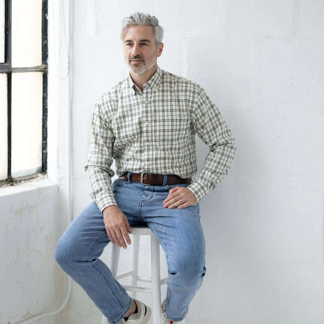 Man wearing a green and black plaid long sleeve button-down shirt, sitting on a stool in a bright, minimalist space.