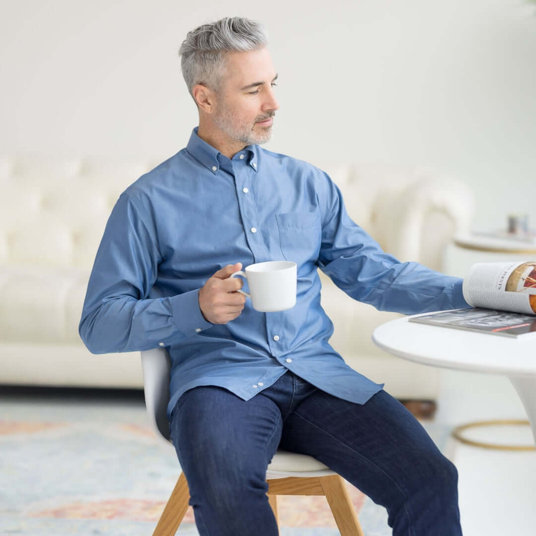 Man in long sleeve blue chambray shirt enjoying coffee and reading, showcasing adaptive fashion with magnetic closures.
