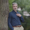 Man in navy long sleeve shirt standing by a tree, showcasing a casual yet stylish look.