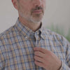 Close-up of a man in a blue and brown plaid long sleeve shirt with button-down collar, adjusting his collar.