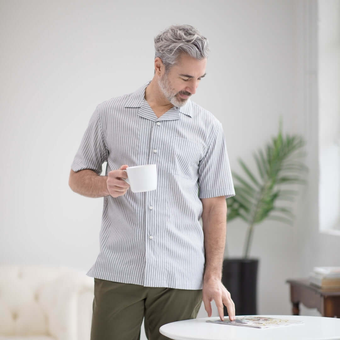 Man in grey short sleeve untucked shirt holding a cup, standing in a bright, modern living space.